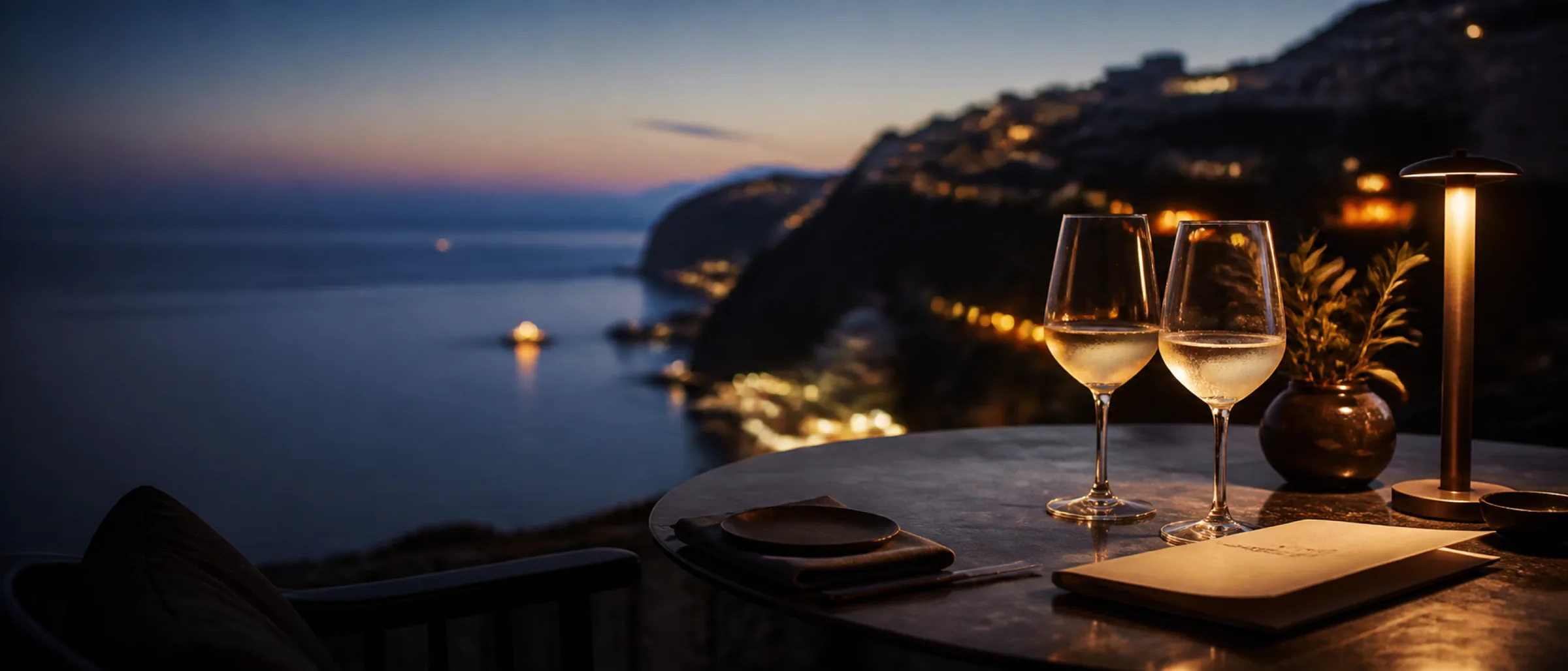 A cliffside terrace table at blue hour on the Mediterranean, two glasses of white wine and a folded menu, a distant harbour out of focus.